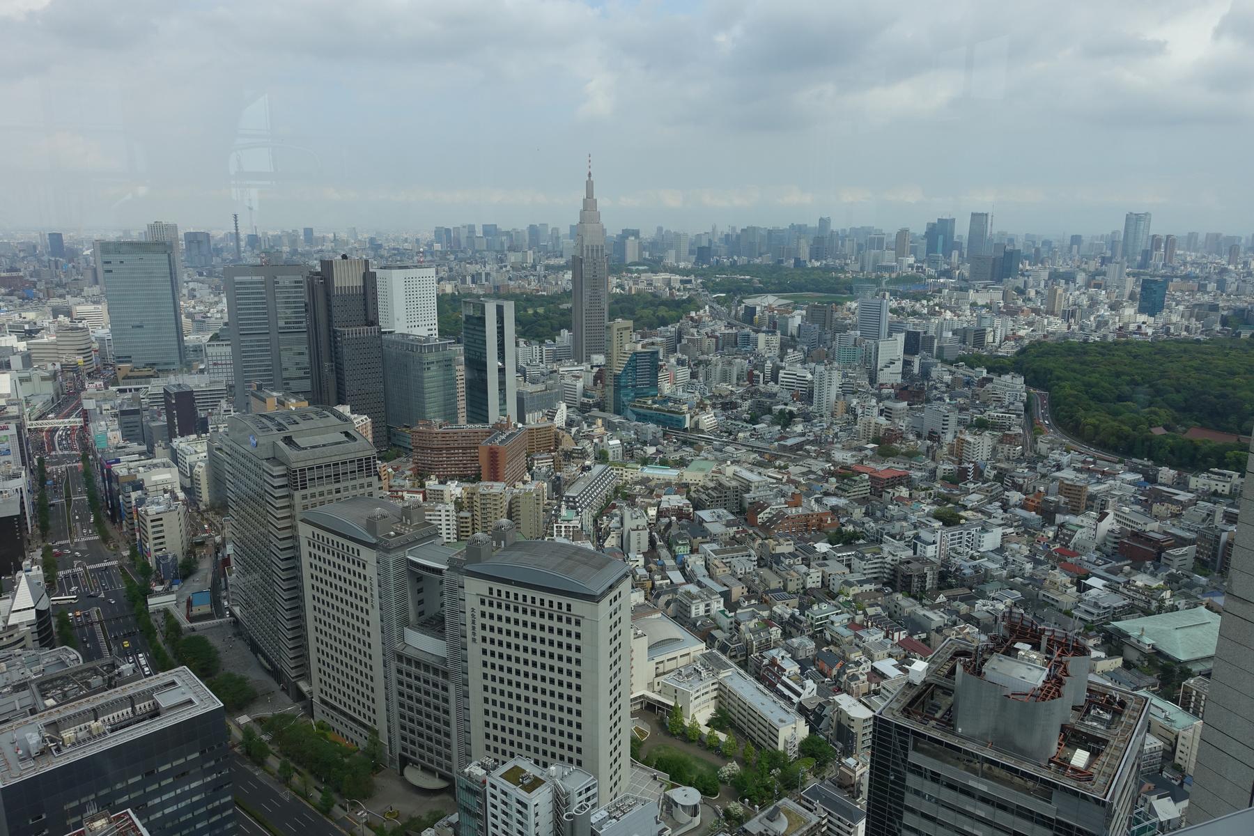A wide view of the Tokyo skyline, featuring many skyscrapers and green spaces under a cloudy sky.