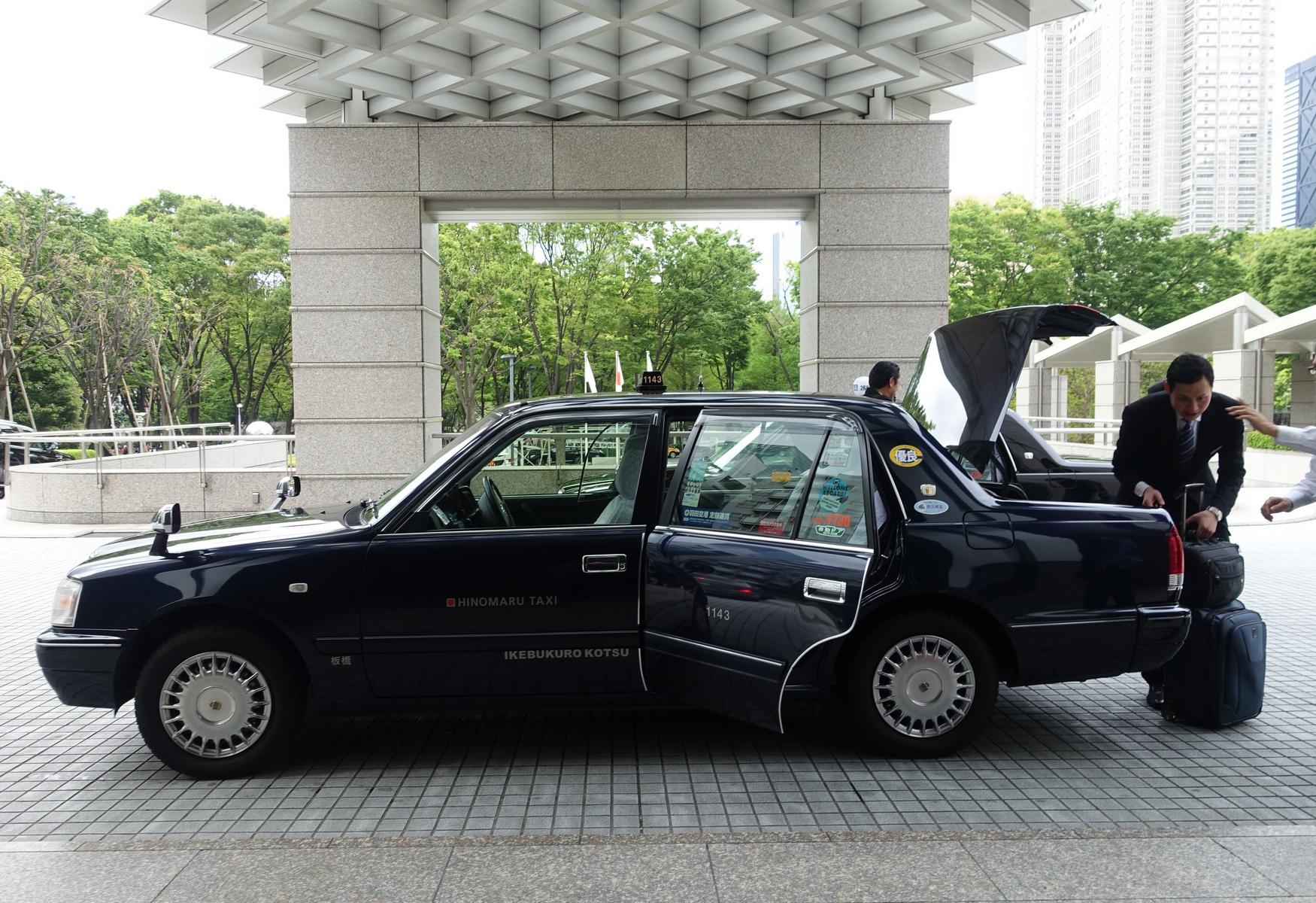 A man loads luggage into a dark Hinomaru taxi at a building entrance.