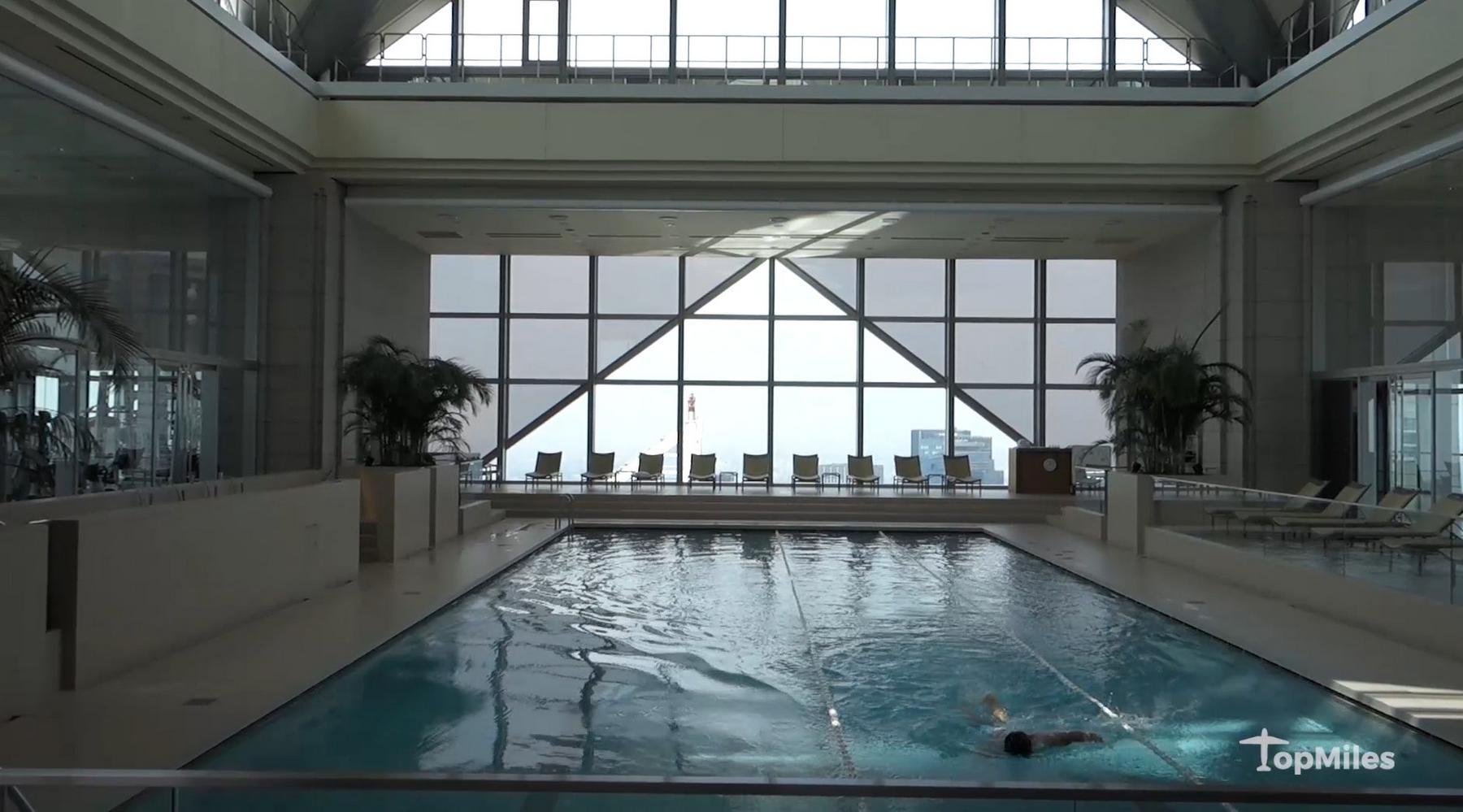 A person swims laps in a luxurious indoor pool with floor-to-ceiling windows overlooking a city view and lounge chairs.