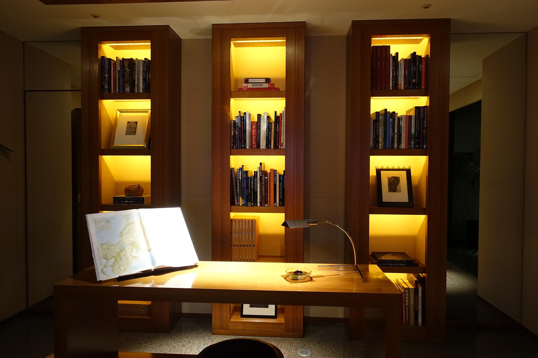 A sophisticated study space featuring a wooden desk with an open atlas and three warmly lit bookshelves filled with books.