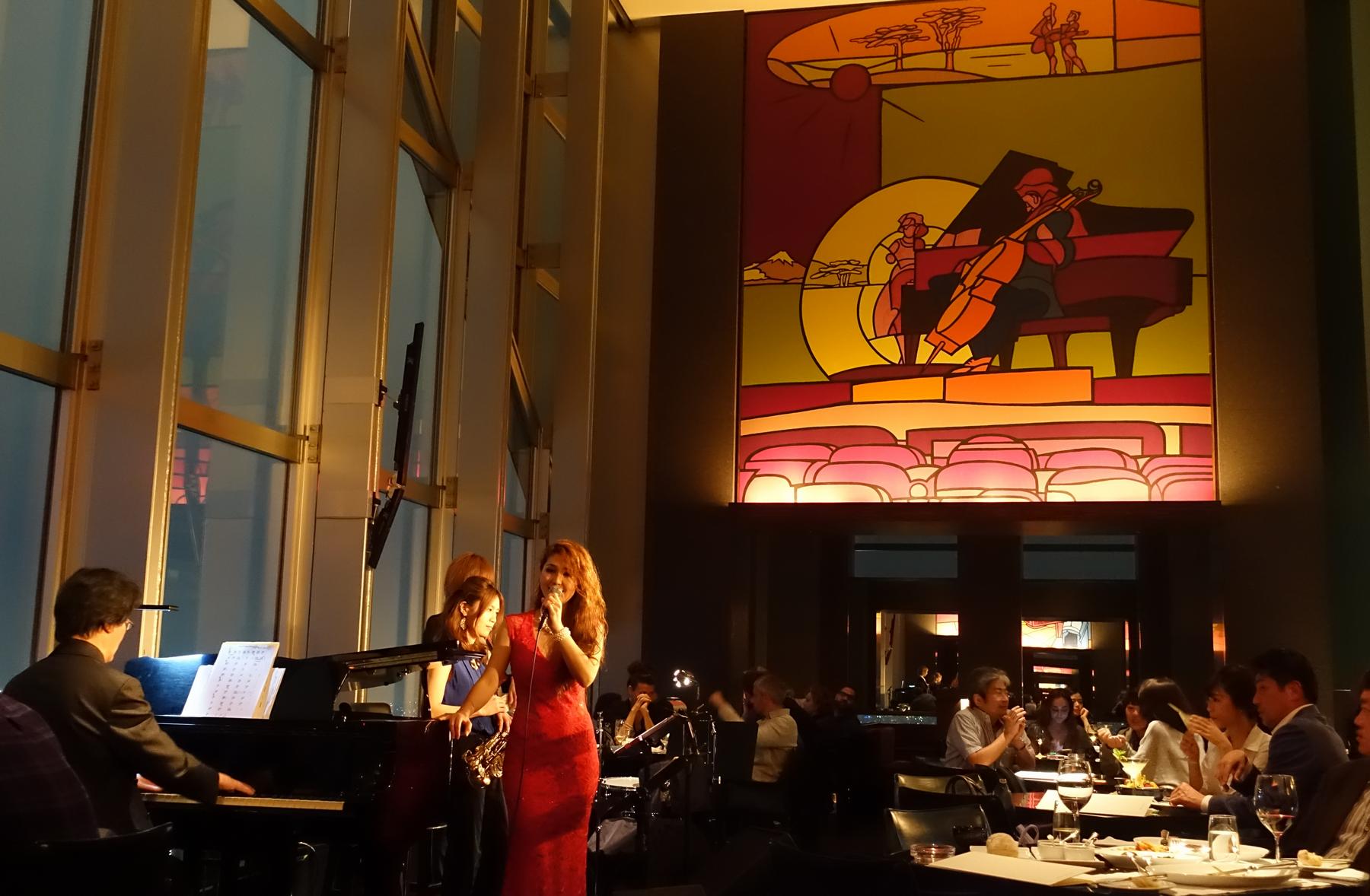 A female singer in a red dress performs with a jazz band for guests in a luxurious bar with a large colorful mural and window views.