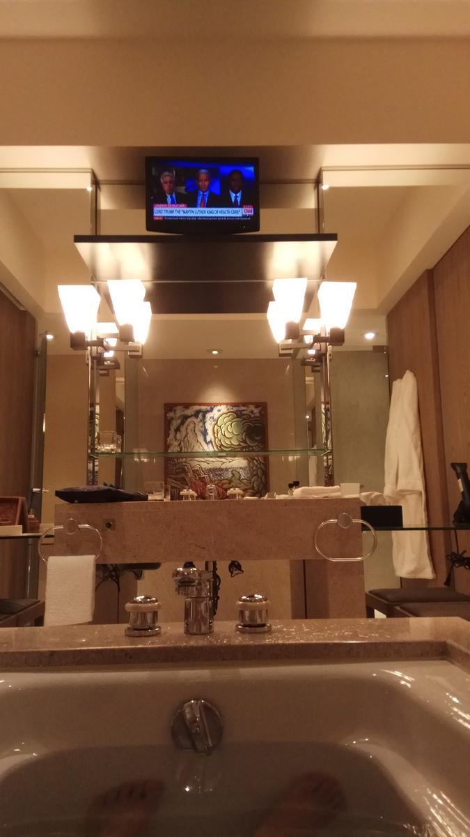 View from a bathtub in a hotel bathroom, showing a news program on a TV mounted above a vanity with mirrors.