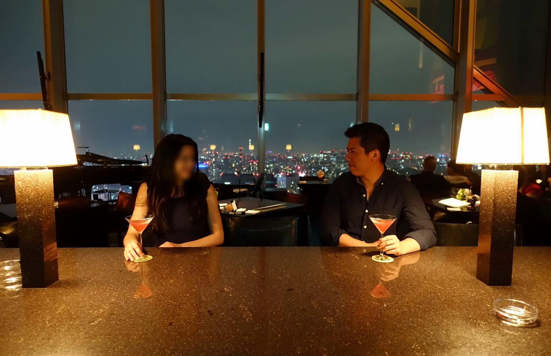 Two people with cocktails sit at a bar overlooking a brightly lit city at night.