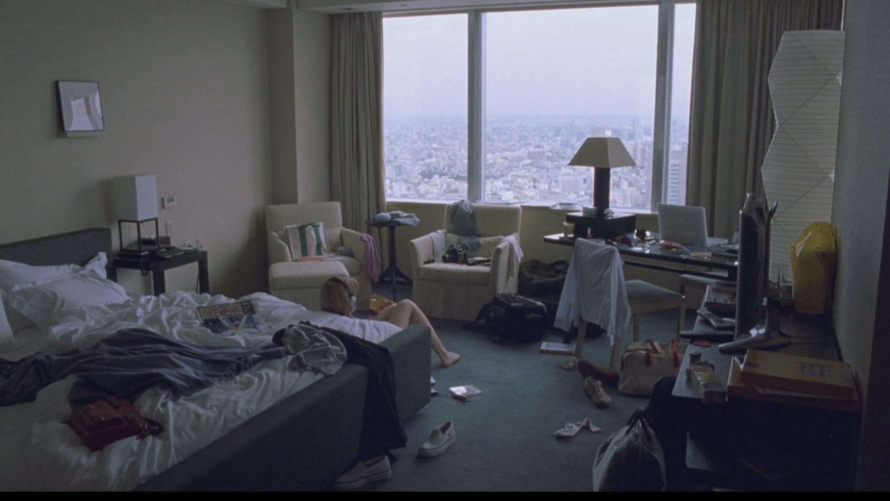 A person lies on the floor of a disheveled hotel room with a large window overlooking a city skyline.