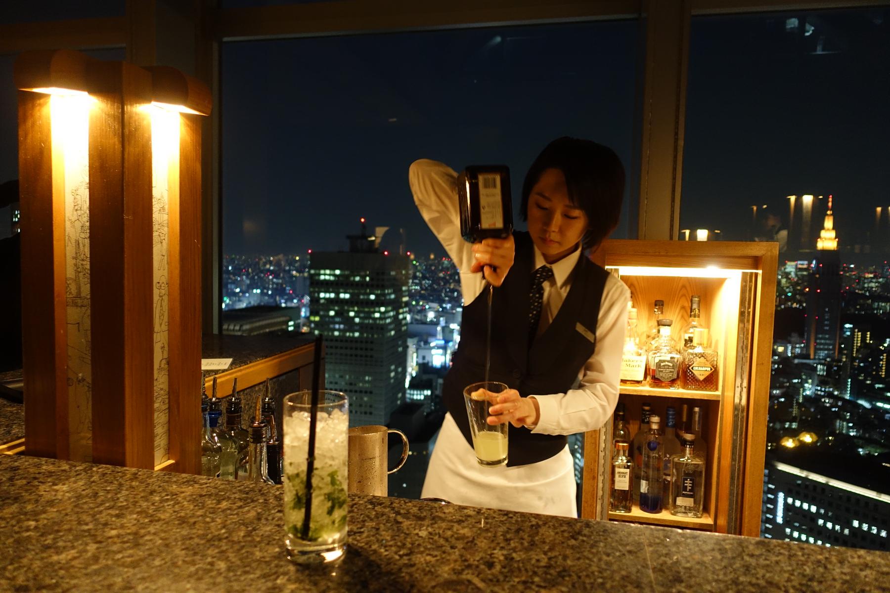 A bartender pours a drink at the Park Hyatt Tokyo's New York Bar, overlooking the city lights at night.