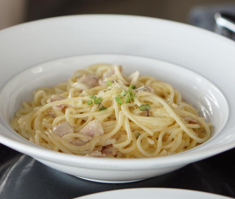 A white bowl of spaghetti carbonara with bacon and microgreens.