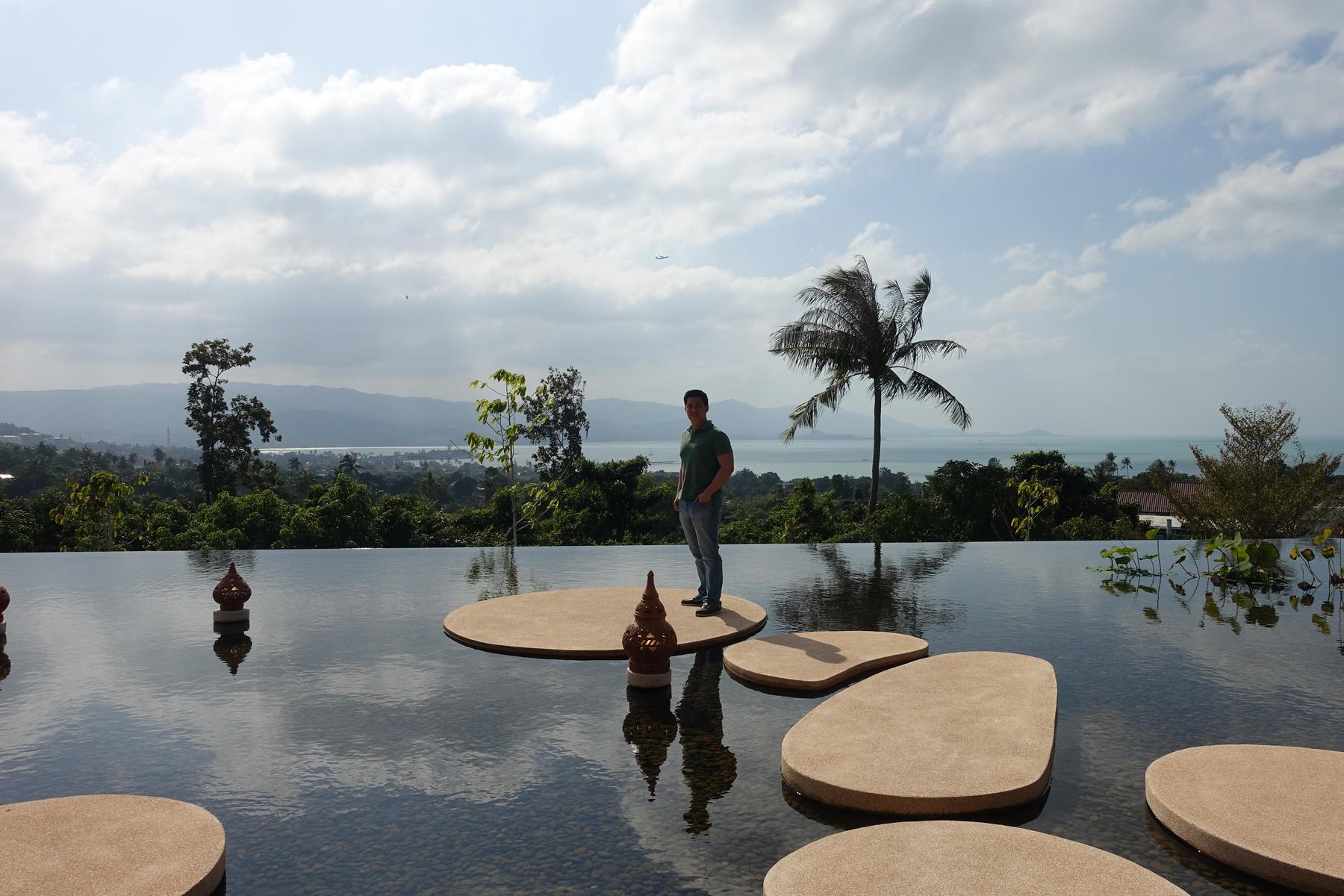 A man stands on a stone platform in an infinity pool overlooking a lush tropical landscape and the ocean.