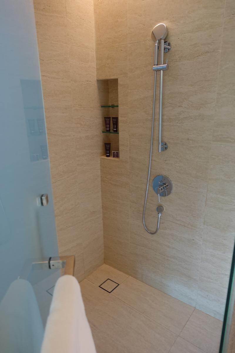 A beige-tiled walk-in shower featuring a handheld showerhead, an inset shelf with toiletries, and a glass door.