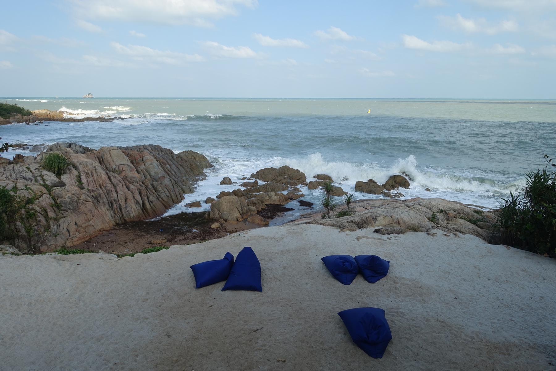 Blue bean bags rest on a sandy beach overlooking a rocky coastline with waves crashing in the ocean.