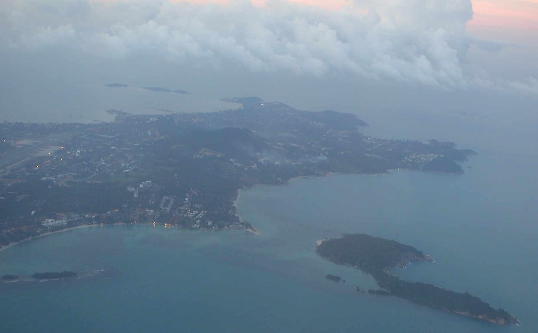 Aerial view of Koh Samui island at dusk, showing coastline, town lights, and clouds.