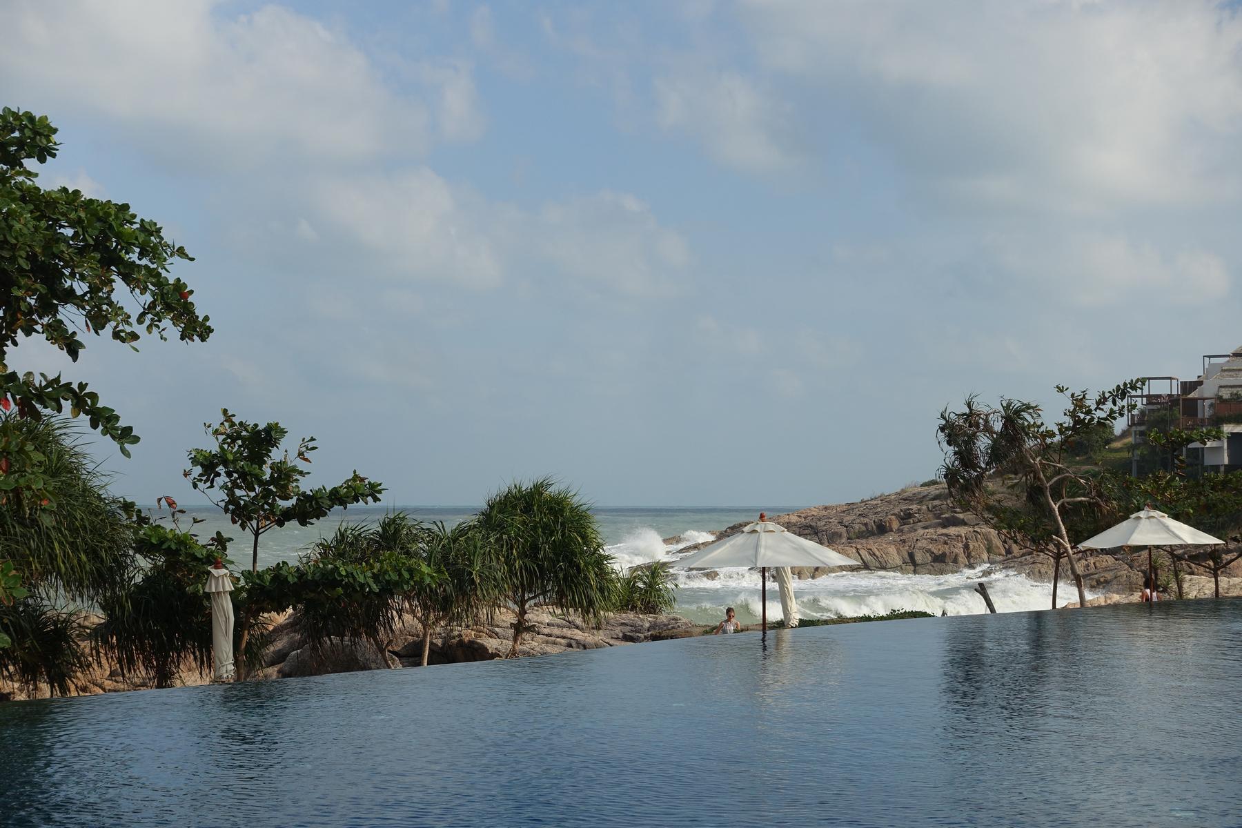An infinity pool overlooks a rocky ocean coastline with crashing waves, tropical trees, and resort buildings.