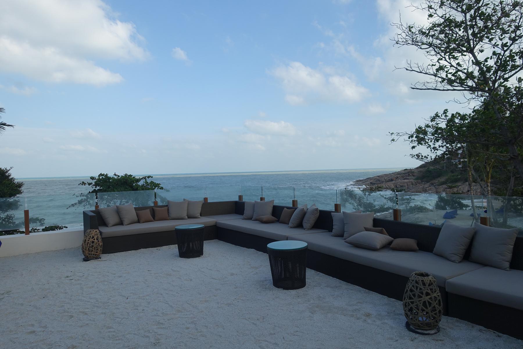 An outdoor lounge with an L-shaped sofa on a sandy patio, overlooking the ocean.