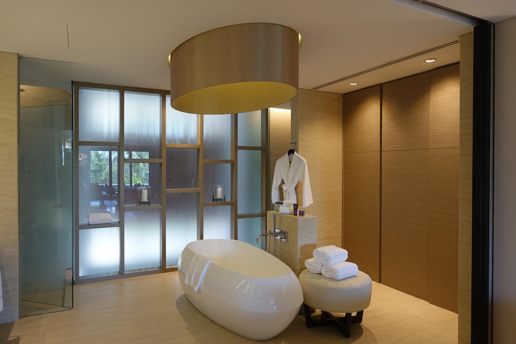 A luxurious modern bathroom with an oval white bathtub, a hanging bathrobe, and a frosted glass wall overlooking tropical trees.