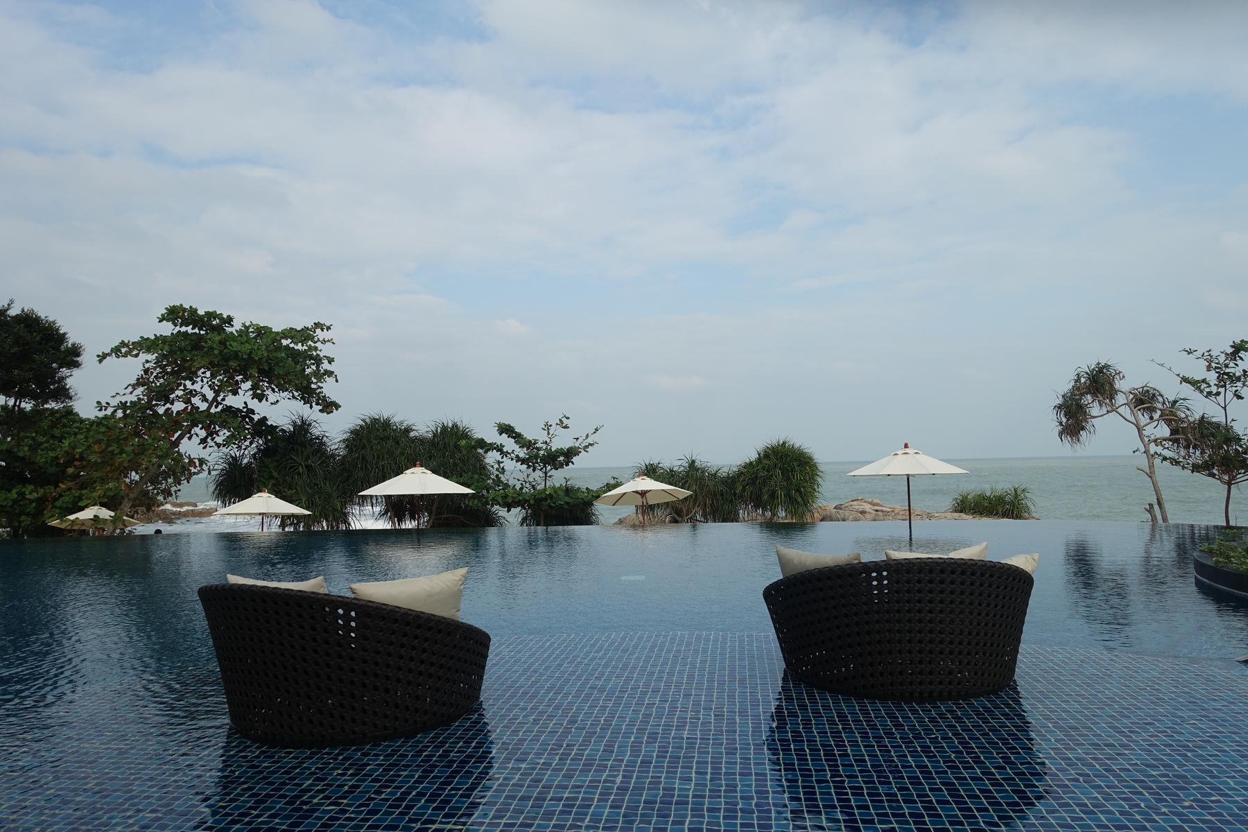 Two woven chairs sit in an infinity pool overlooking the ocean, tropical trees, and beach umbrellas.