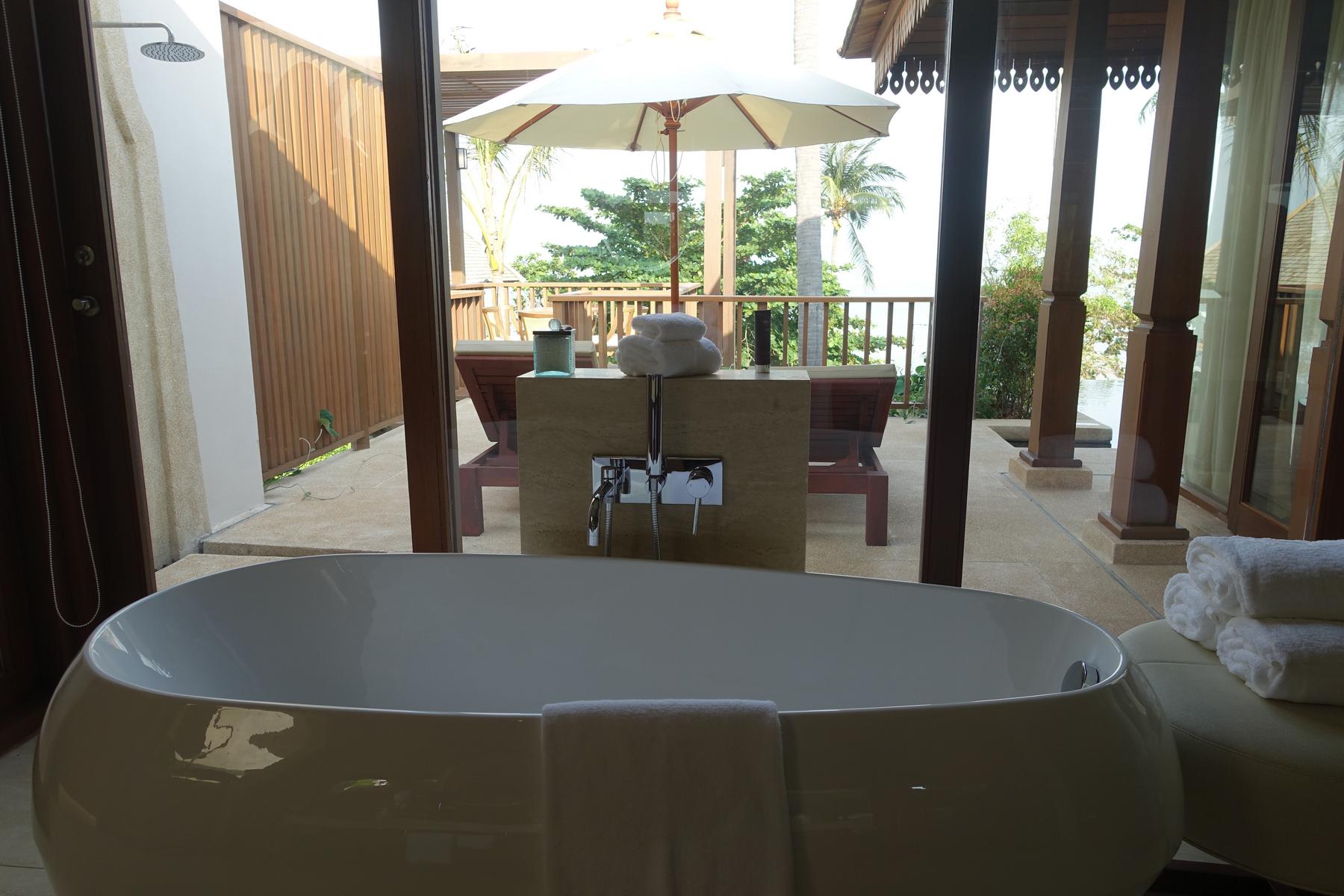 A white freestanding bathtub facing a glass wall that opens to an outdoor patio with two lounge chairs and tropical plants.