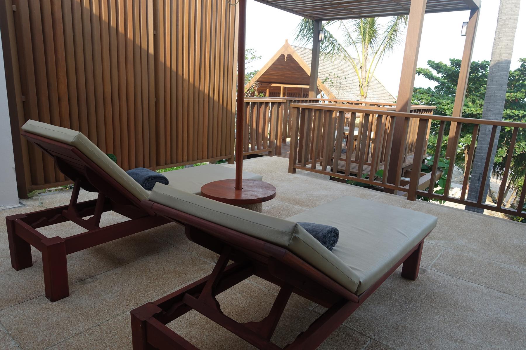 An outdoor patio features two lounge chairs with towels, a small table, wooden railings, and palm trees in the background.