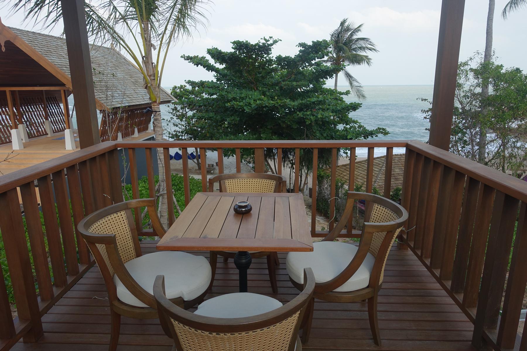A wooden balcony with a table and chairs overlooks the ocean and palm trees.