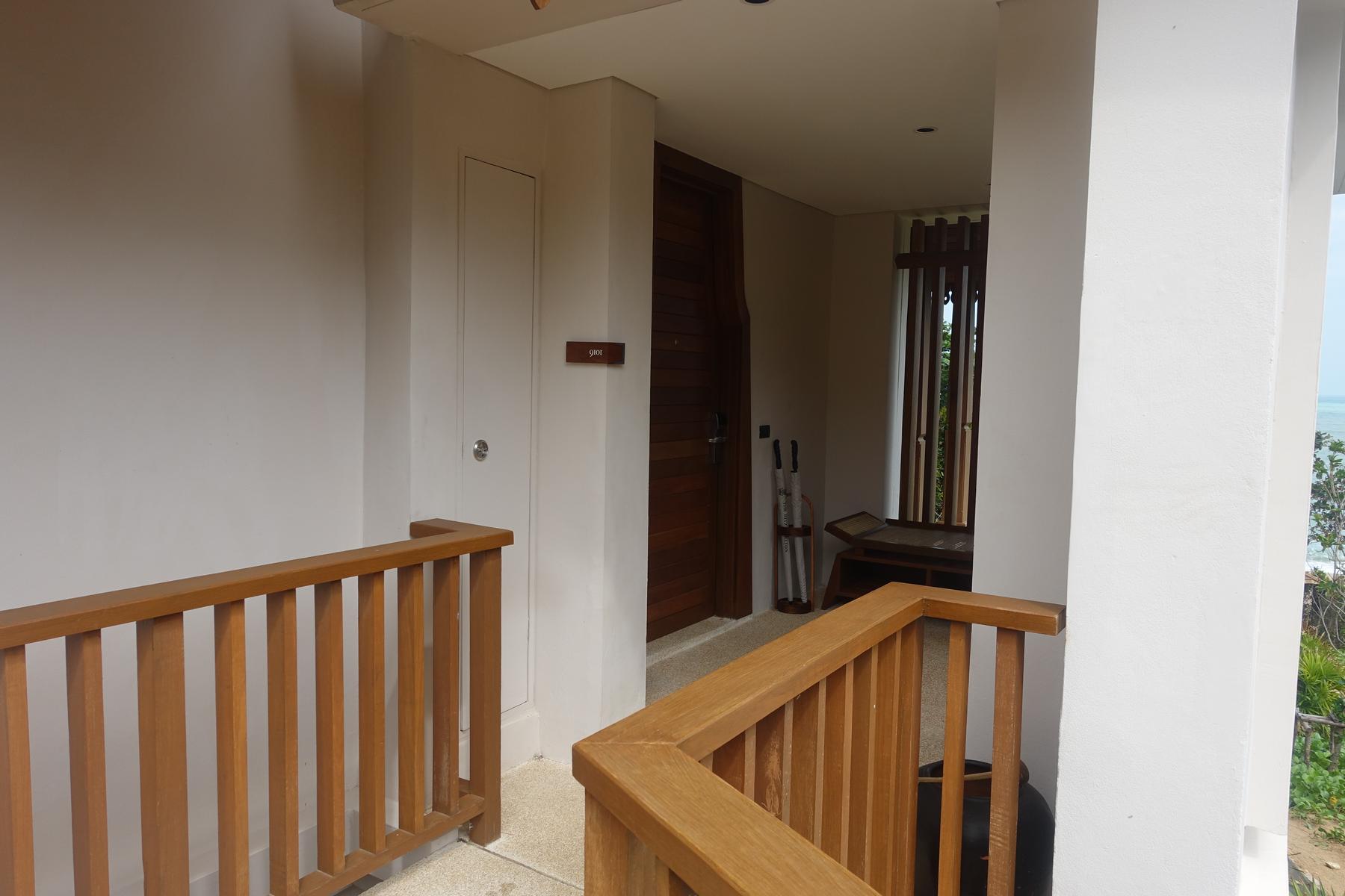 A resort hallway with wooden railings leads to room 9101, showing a partial ocean view.