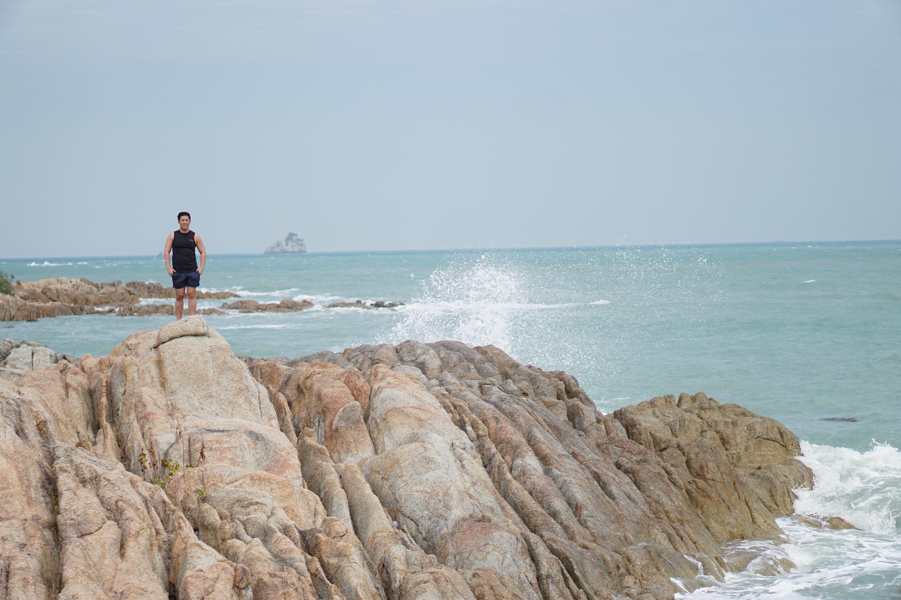 A man stands on a rocky coast with ocean waves crashing and an island in the distance.