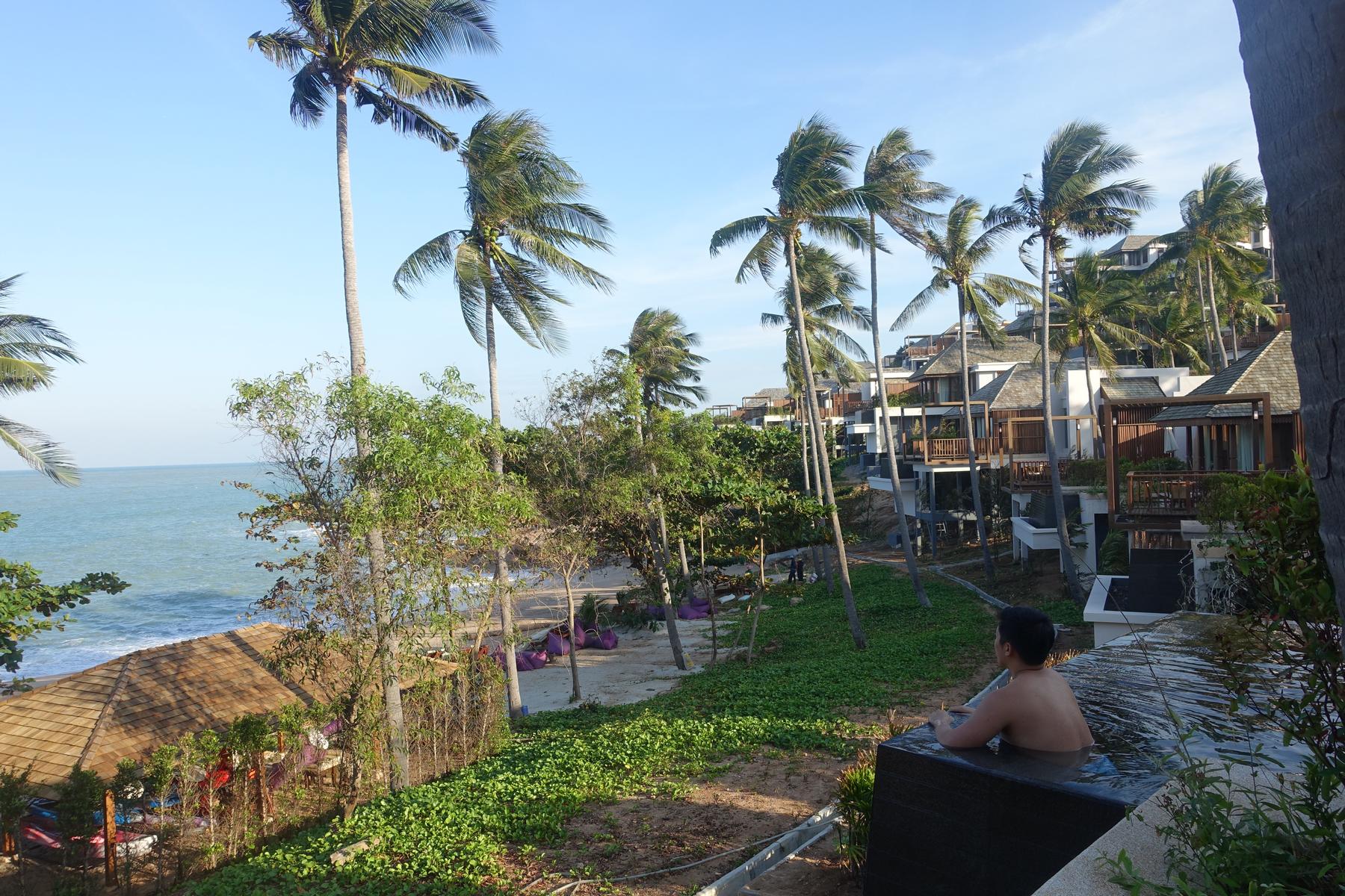 A person relaxing in a private pool with a view of the beach, palm trees, and resort villas.