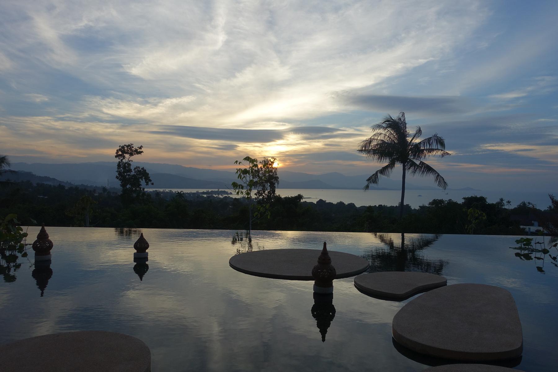 An infinity pool with decorative stones reflects a vibrant sunset over a tropical bay and mountains.