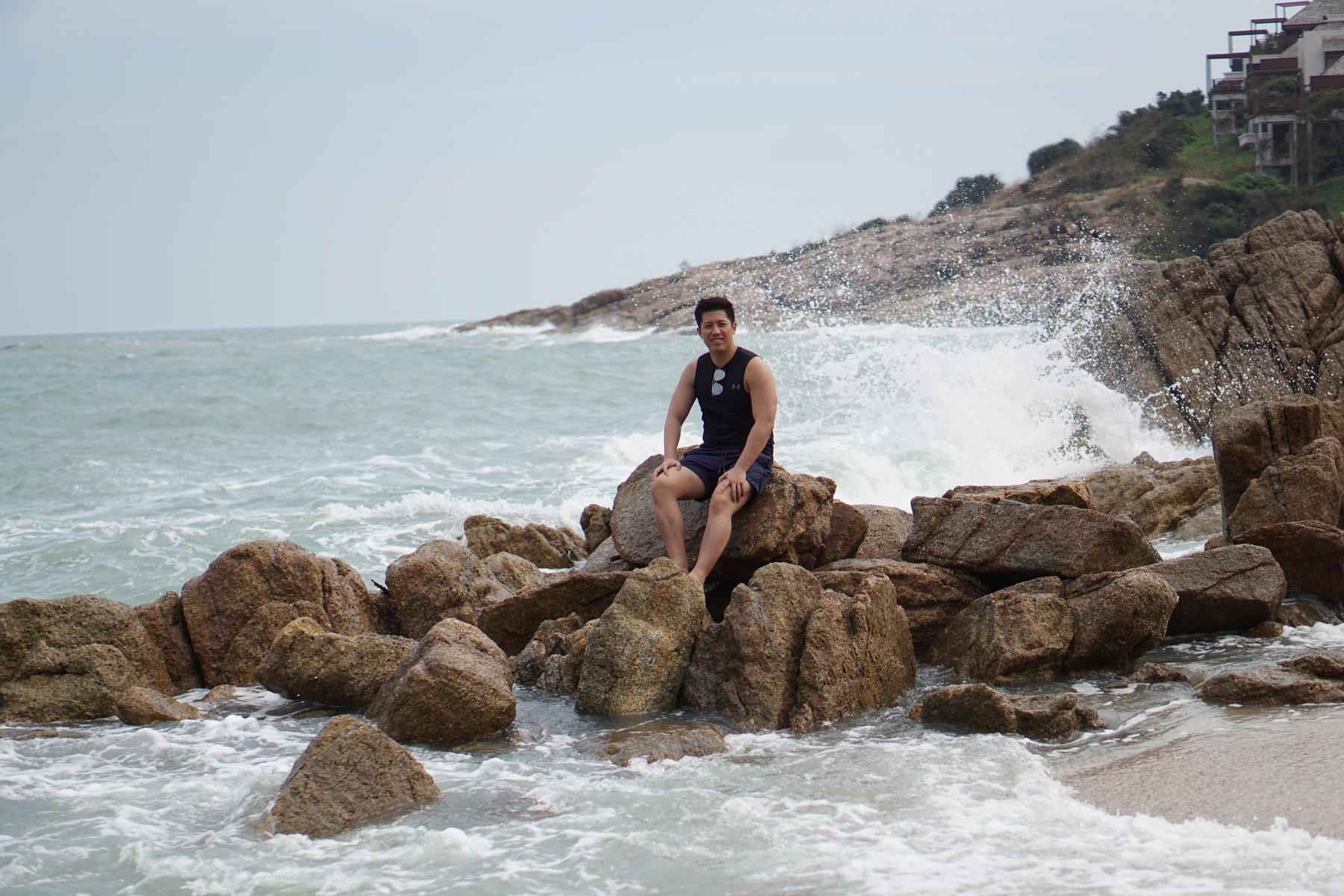 A man sits smiling on rocks at a beach with a large wave crashing behind him.