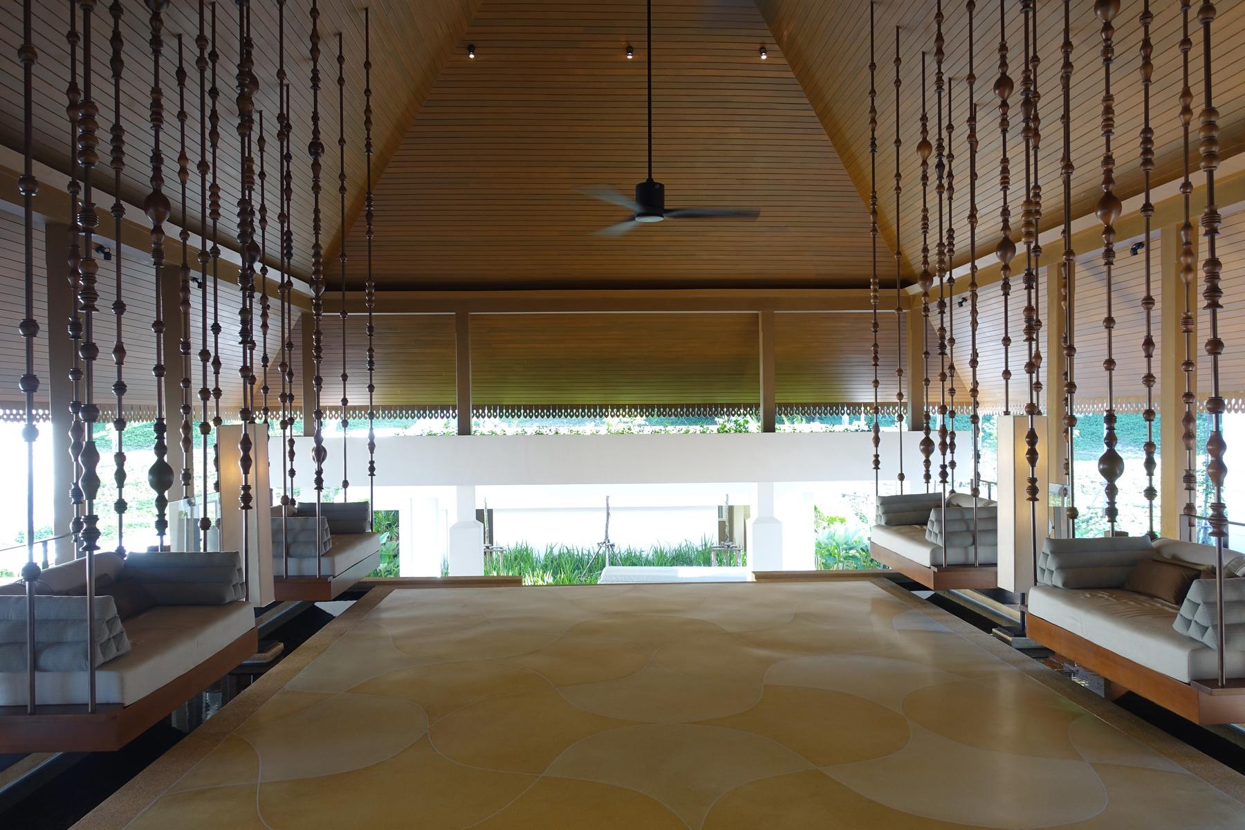 A spacious, open-air resort lobby with a wooden ceiling, patterned floor, and suspended daybeds.