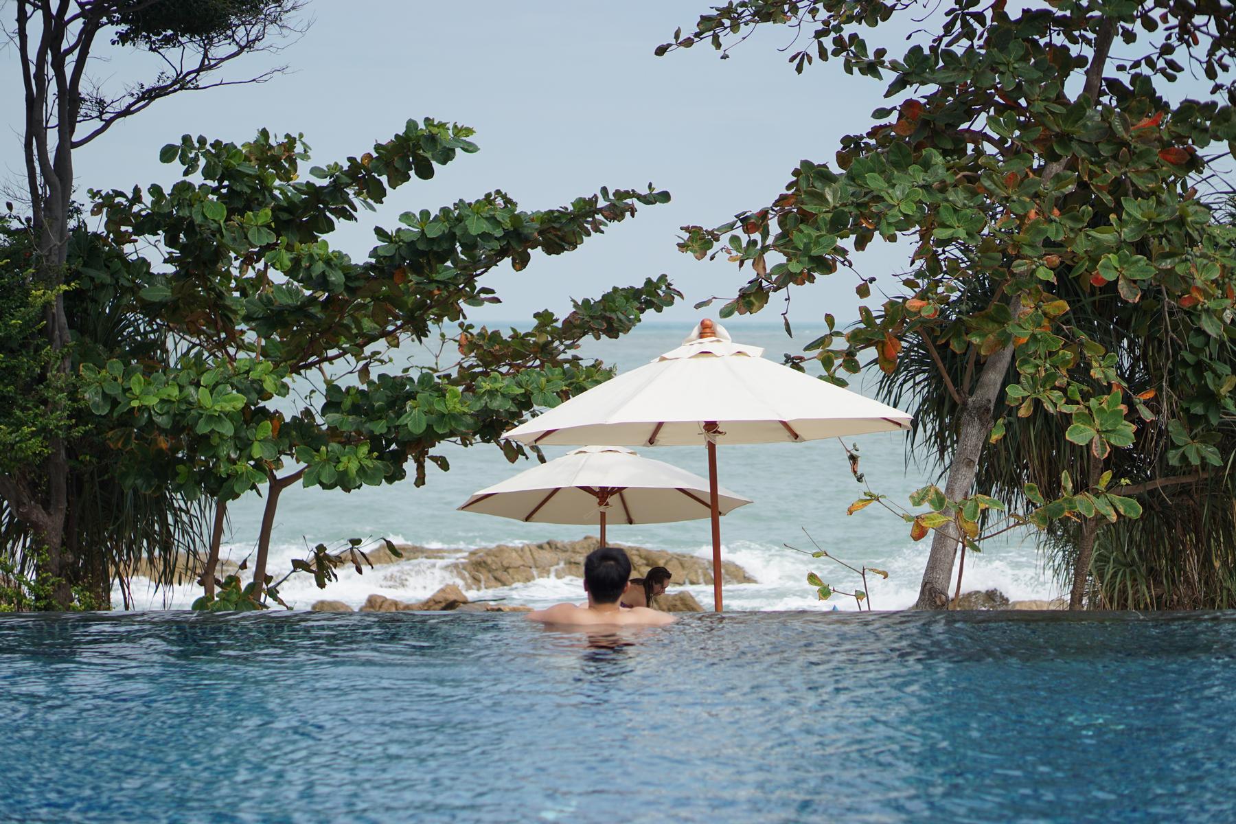 Two people in an infinity pool overlooking the ocean with beach umbrellas and tropical trees.