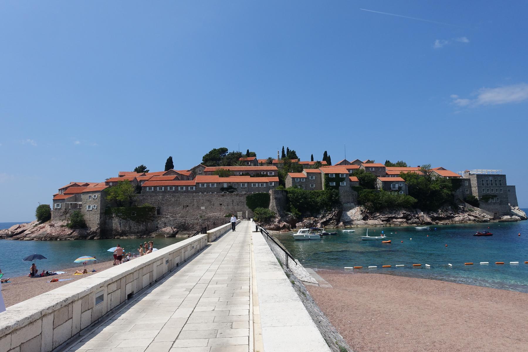 A stone causeway connects a sandy beach to Sveti Stefan islet, featuring historic stone buildings with red roofs, surrounded by clear blue water under a sunny sky.