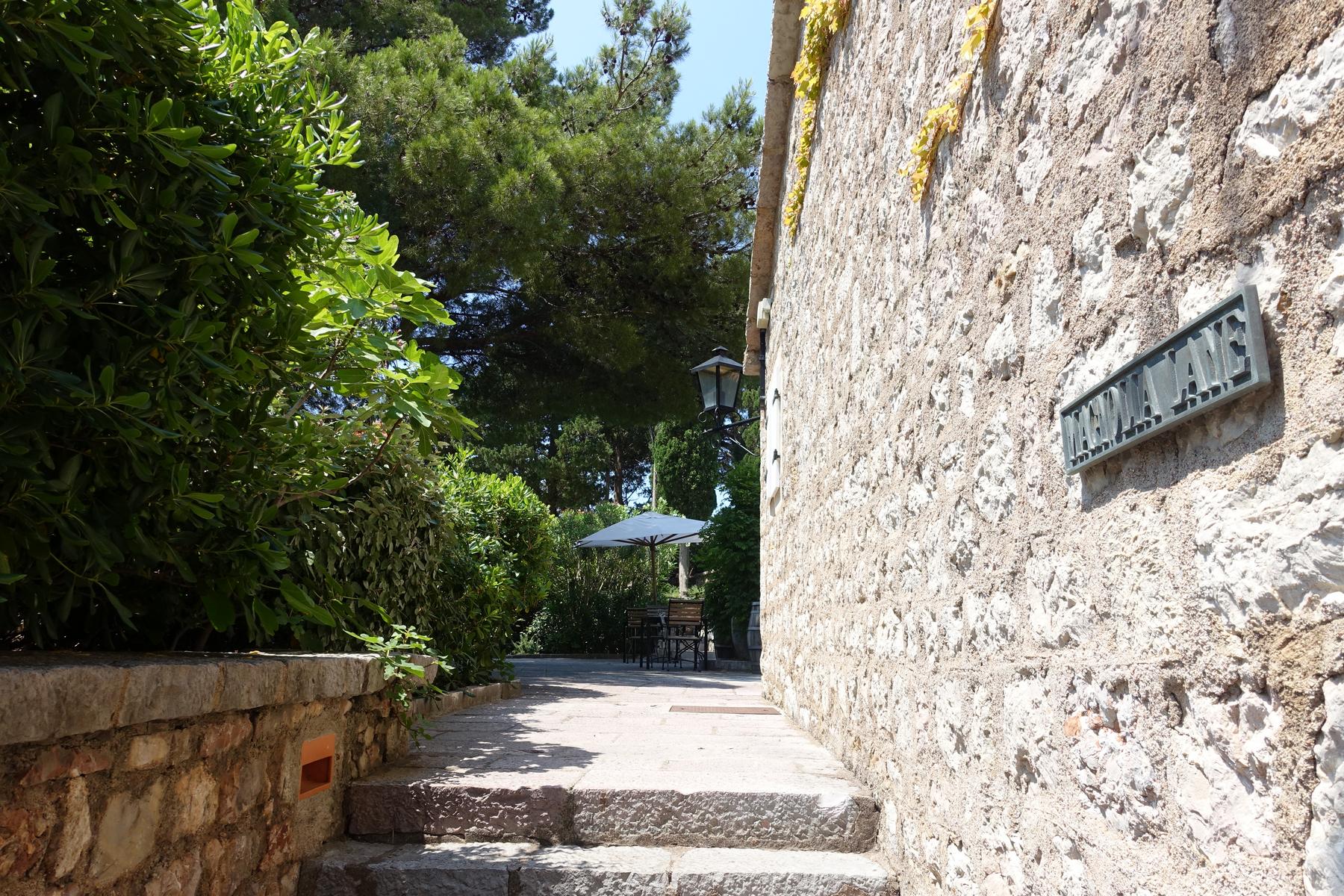 Stone steps lead along a textured stone wall with a "MADIA LANE" sign and lush green foliage, towards an outdoor seating area with an umbrella.