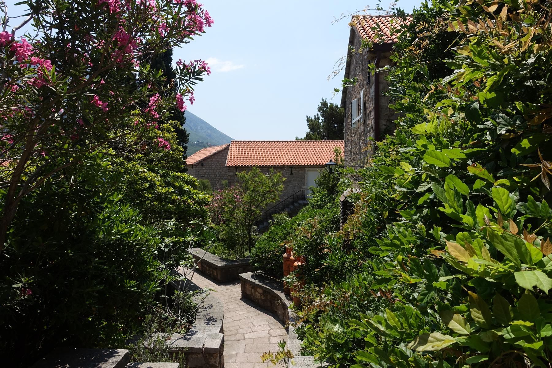 A winding stone path through lush green foliage and pink flowers leads to traditional stone buildings with orange roofs.