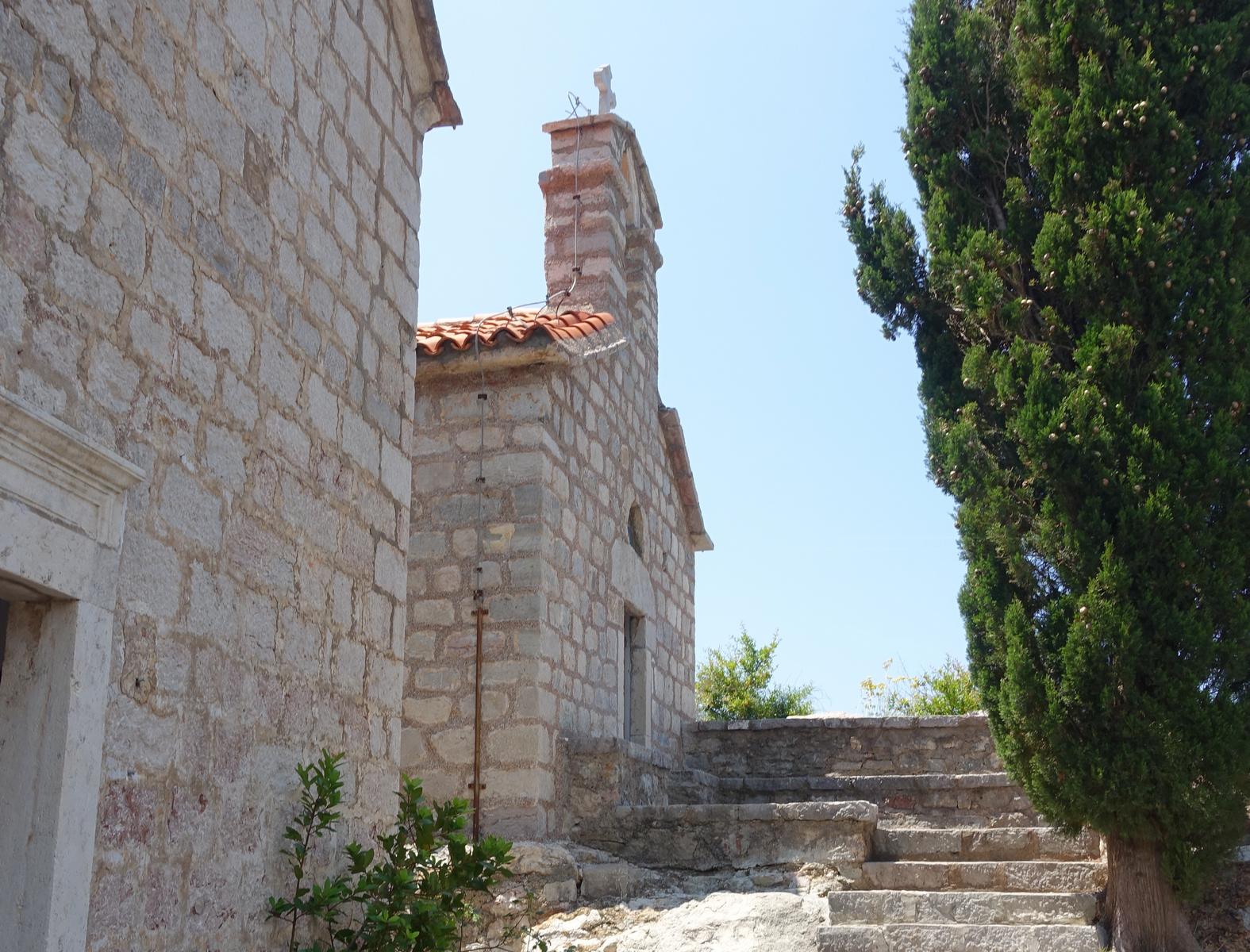 Stone buildings with terracotta roofs and steps, next to a tall cypress tree against a clear blue sky on Sveti Stefan.