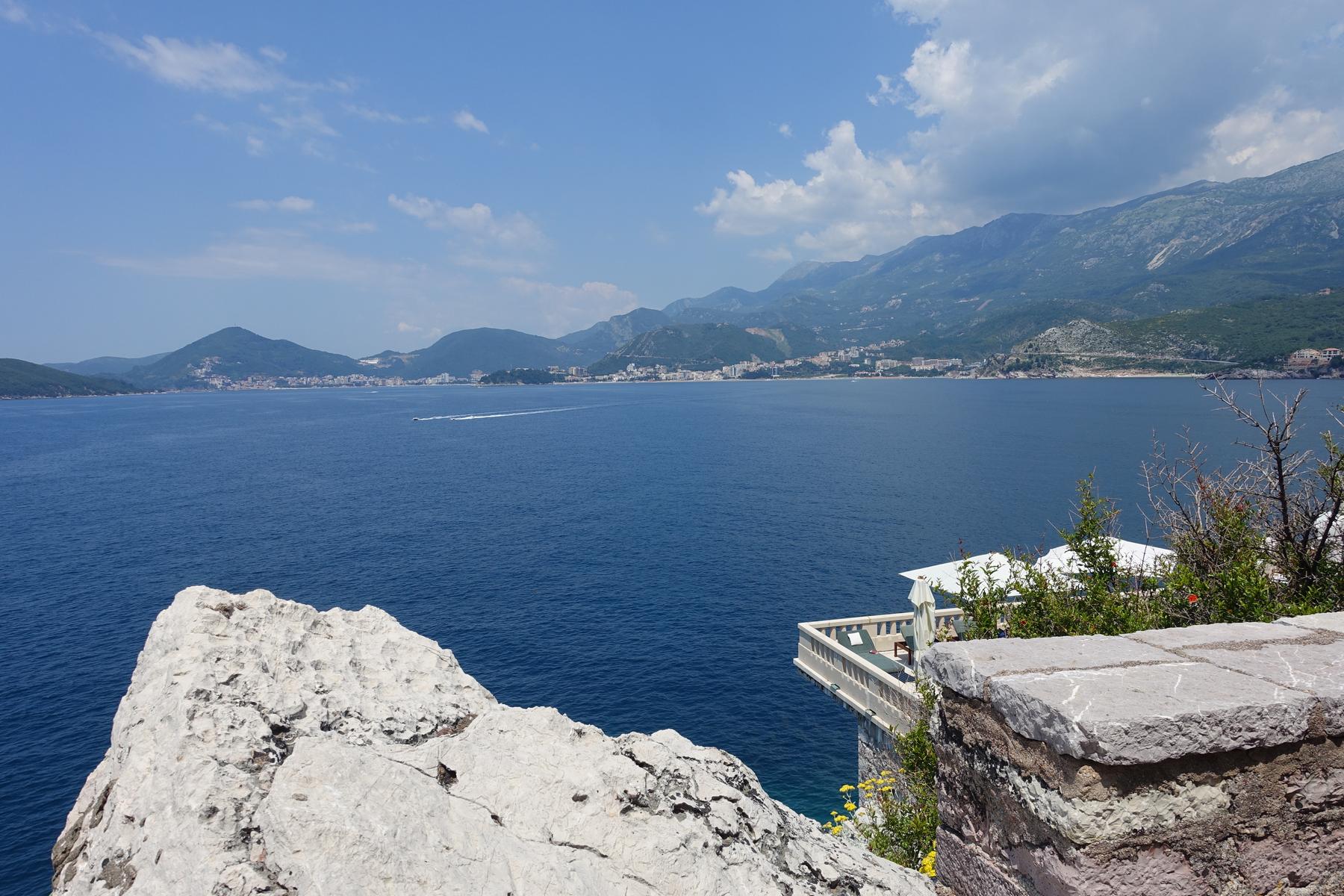 Panoramic view of the deep blue sea, a distant mountainous coastline with towns, and a luxurious cliffside balcony in the foreground.