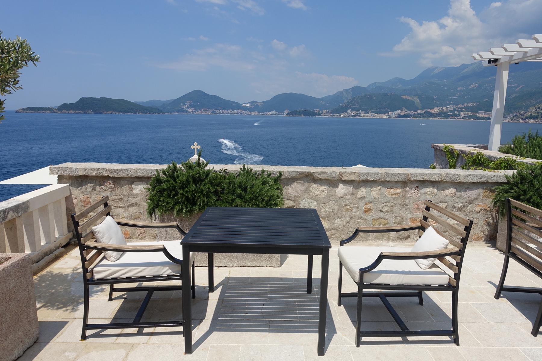 Outdoor seating on a patio offers a scenic view of the blue sea and distant mountainous coastline.