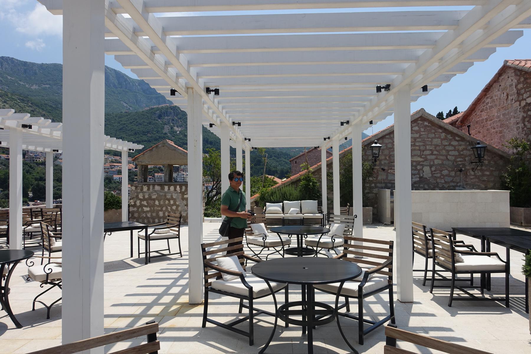 A man stands on a sunlit outdoor terrace with white pergolas, tables, and a mountain backdrop.