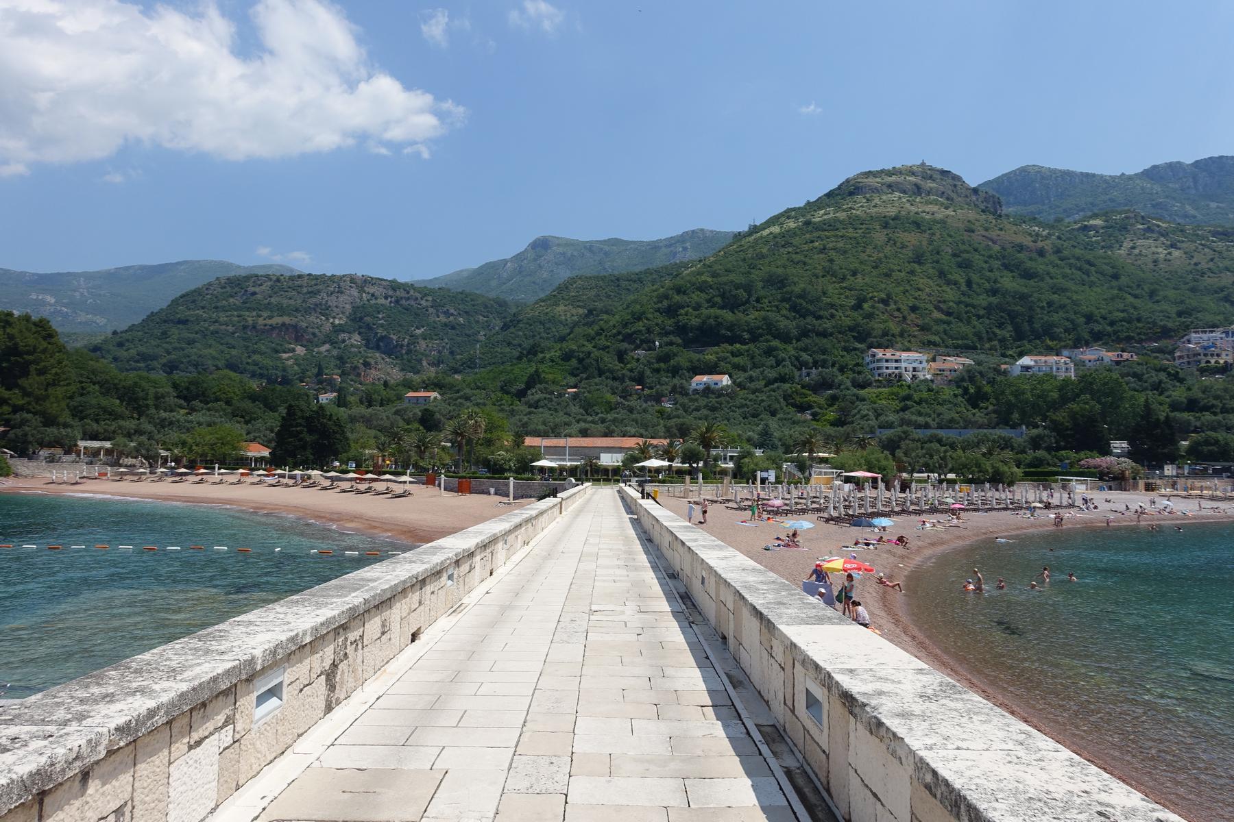The Sveti Stefan causeway extends toward forested mountains, flanked by two sandy beaches bustling with swimmers and sunbathers.