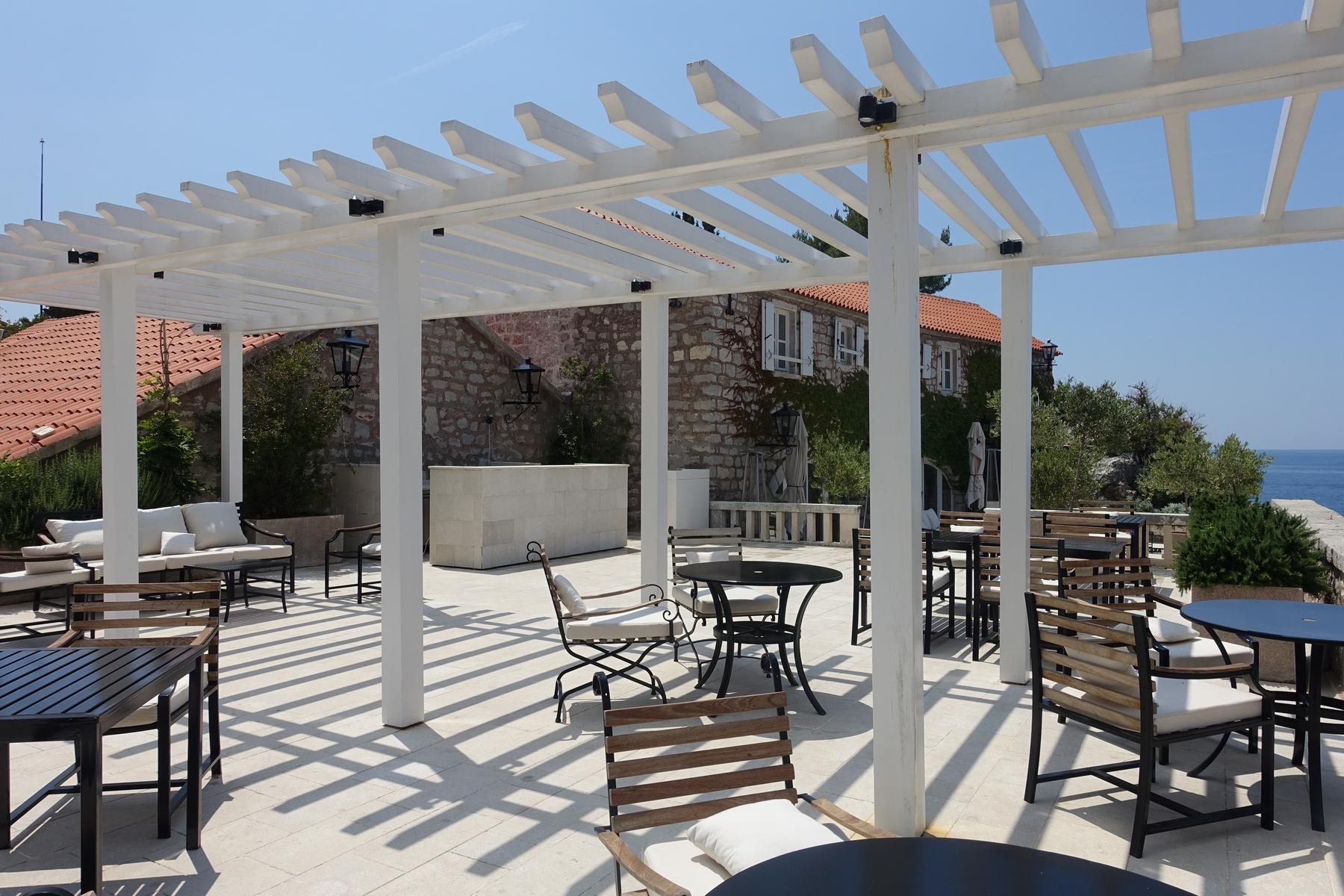 A sunny outdoor terrace at Sveti Stefan with a white pergola, elegant tables and chairs, and a stone building, overlooking the blue sea.