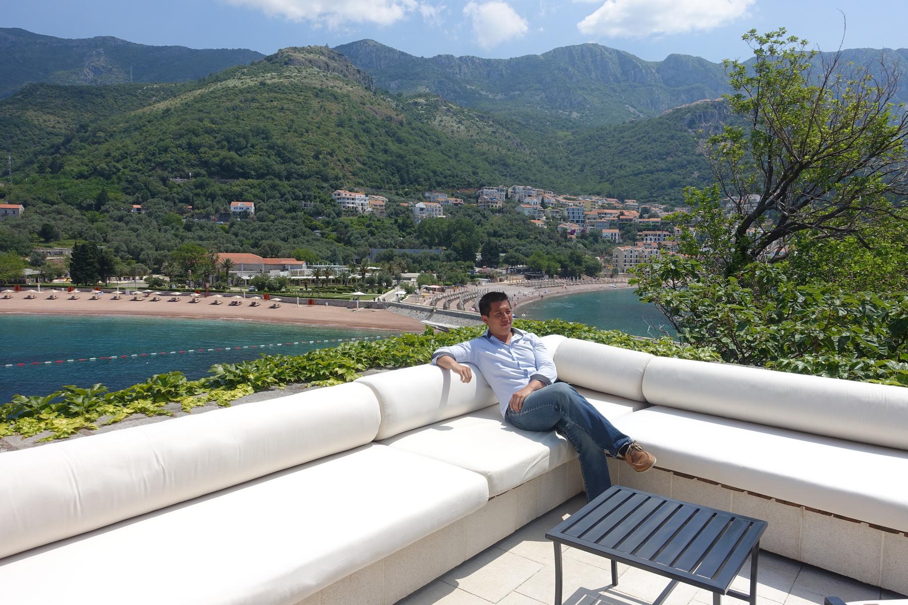 A man relaxes on a terrace sofa overlooking Sveti Stefan bay with its beach and mountains.