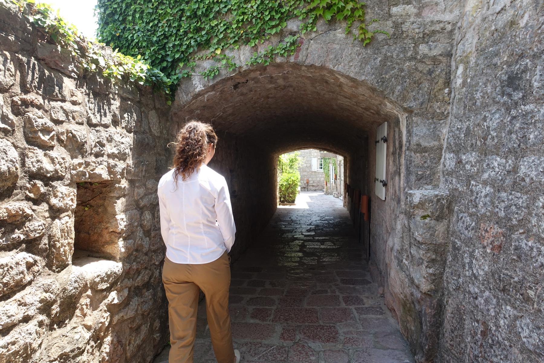 A person looks out from a dark stone archway to a bright sunlit path.