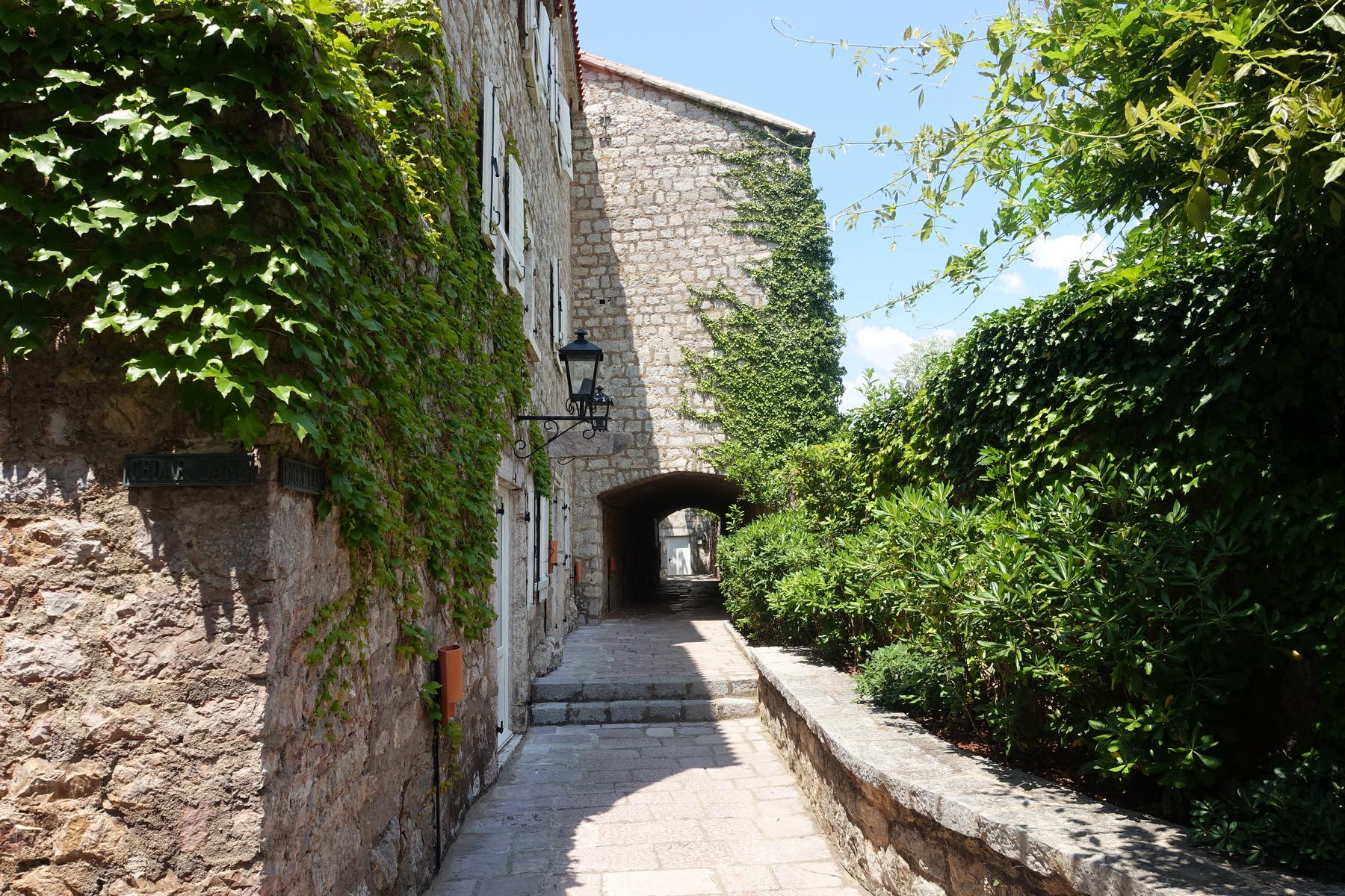 A narrow stone alleyway lined with ivy-covered buildings and lush greenery, leading to an arched passage.