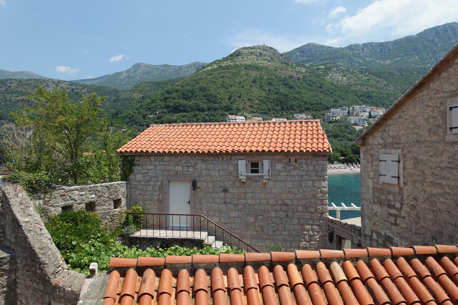 Stone buildings with red tile roofs overlook the sea and green mountains.