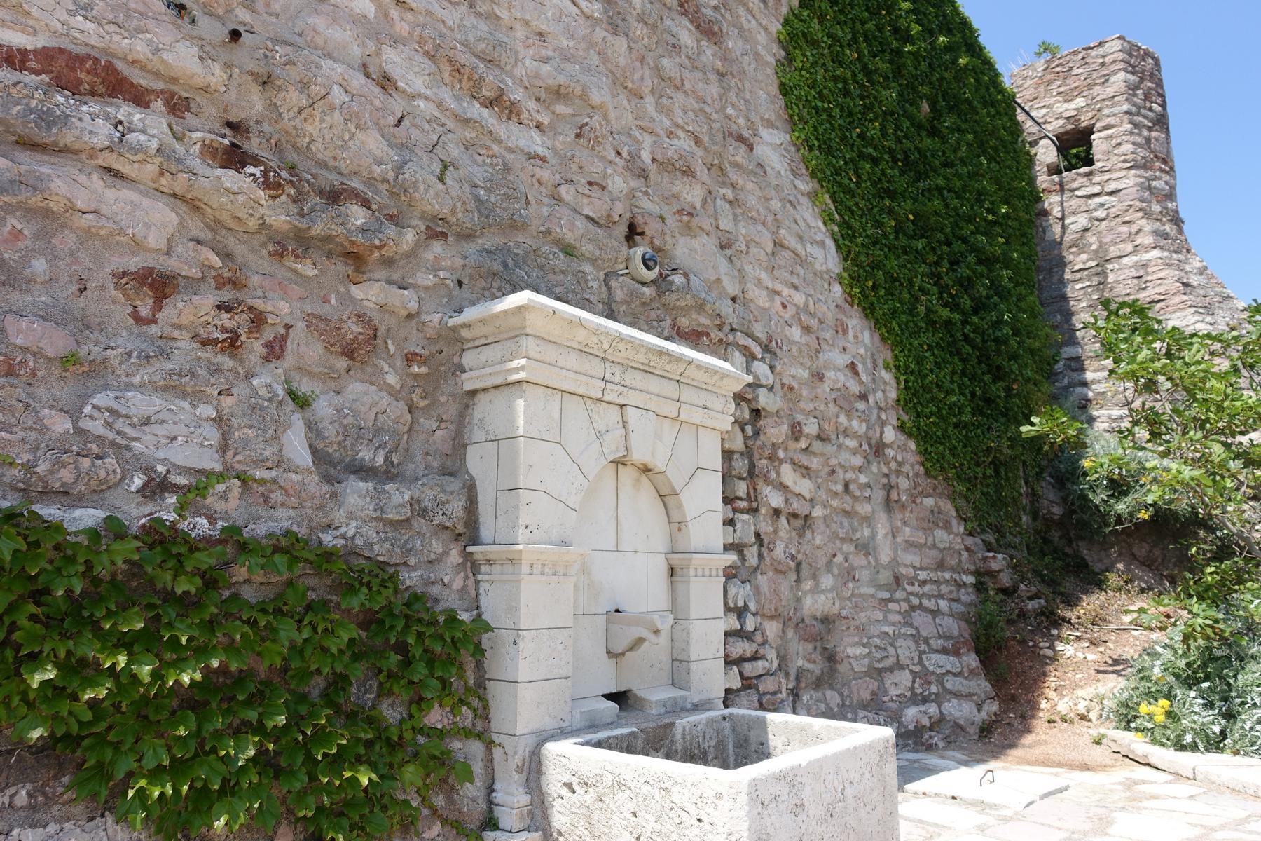 A decorative stone fountain is set into a rustic stone wall covered with green ivy, with a stone tower in the background.