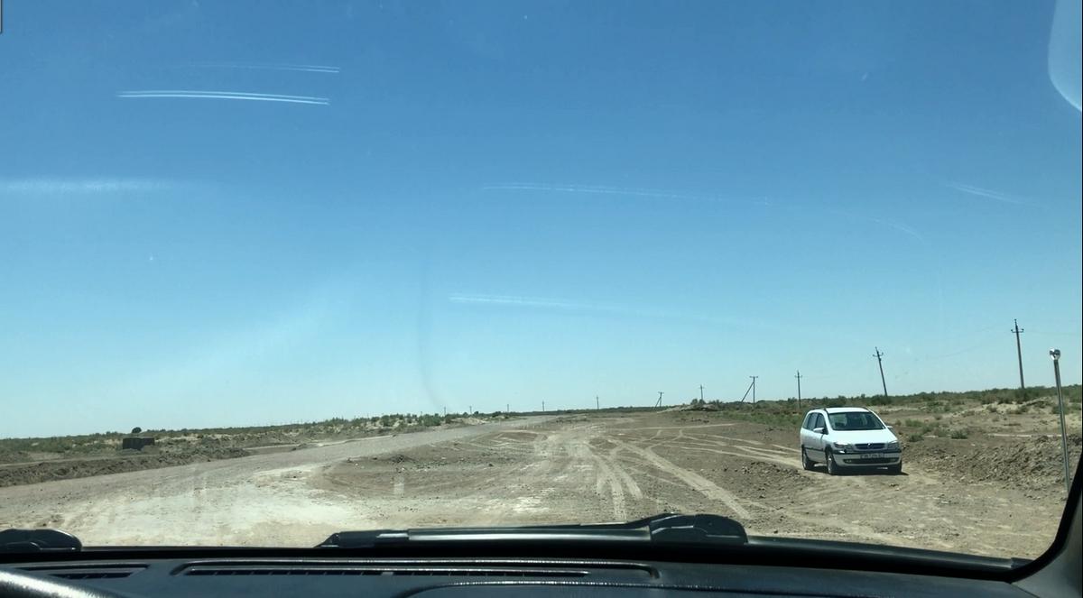View from a car dashboard showing a white van on a wide dirt track in a desert landscape under a clear blue sky.