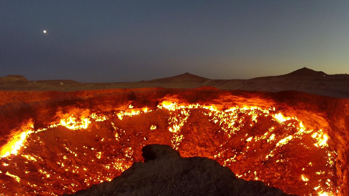 The Darvaza Gas Crater, a large fiery pit, glows under a dark sky with a bright moon.