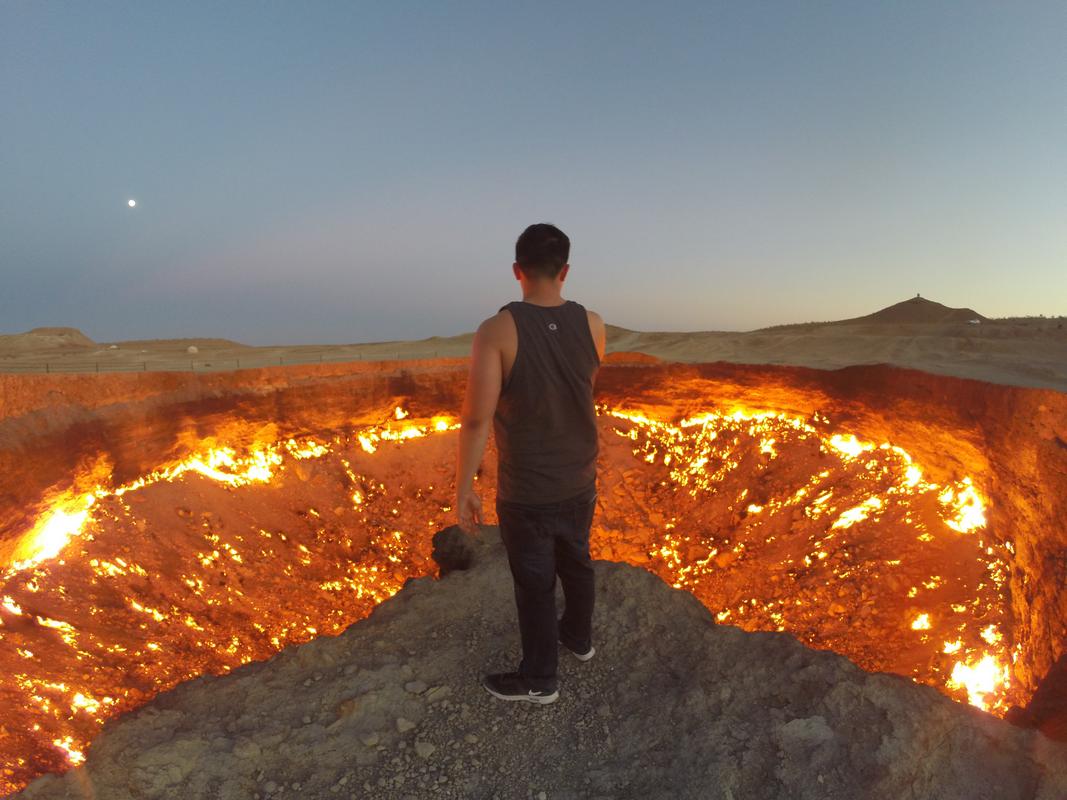 A person stands at the edge of the immense, fiery Darvaza Gas Crater at dusk.