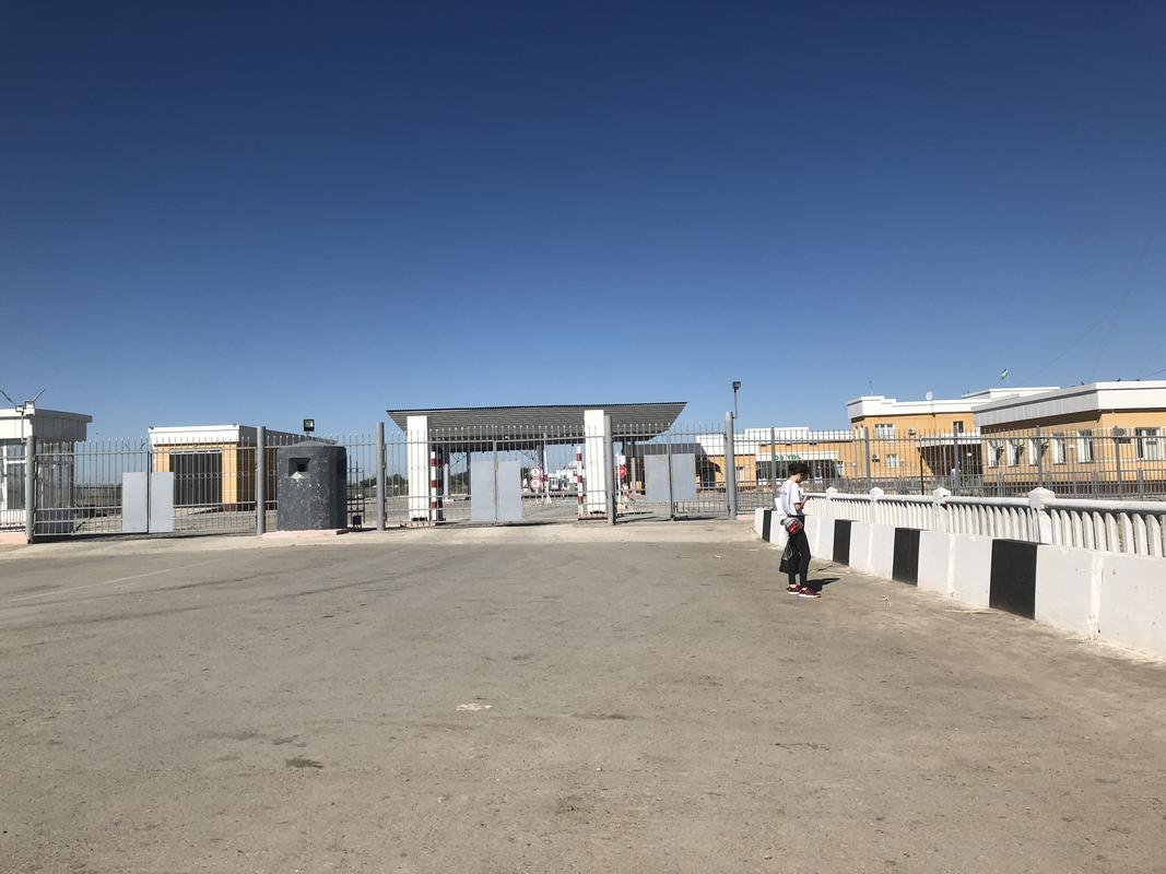 A person stands on a paved area in front of a fenced checkpoint with buildings under a clear blue sky.