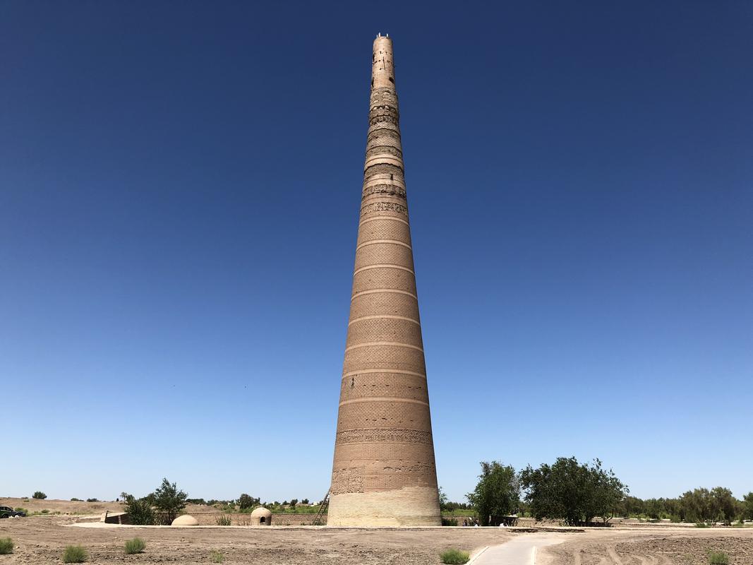 A tall, tapering brick minaret with decorative bands stands against a clear blue sky in an arid landscape.