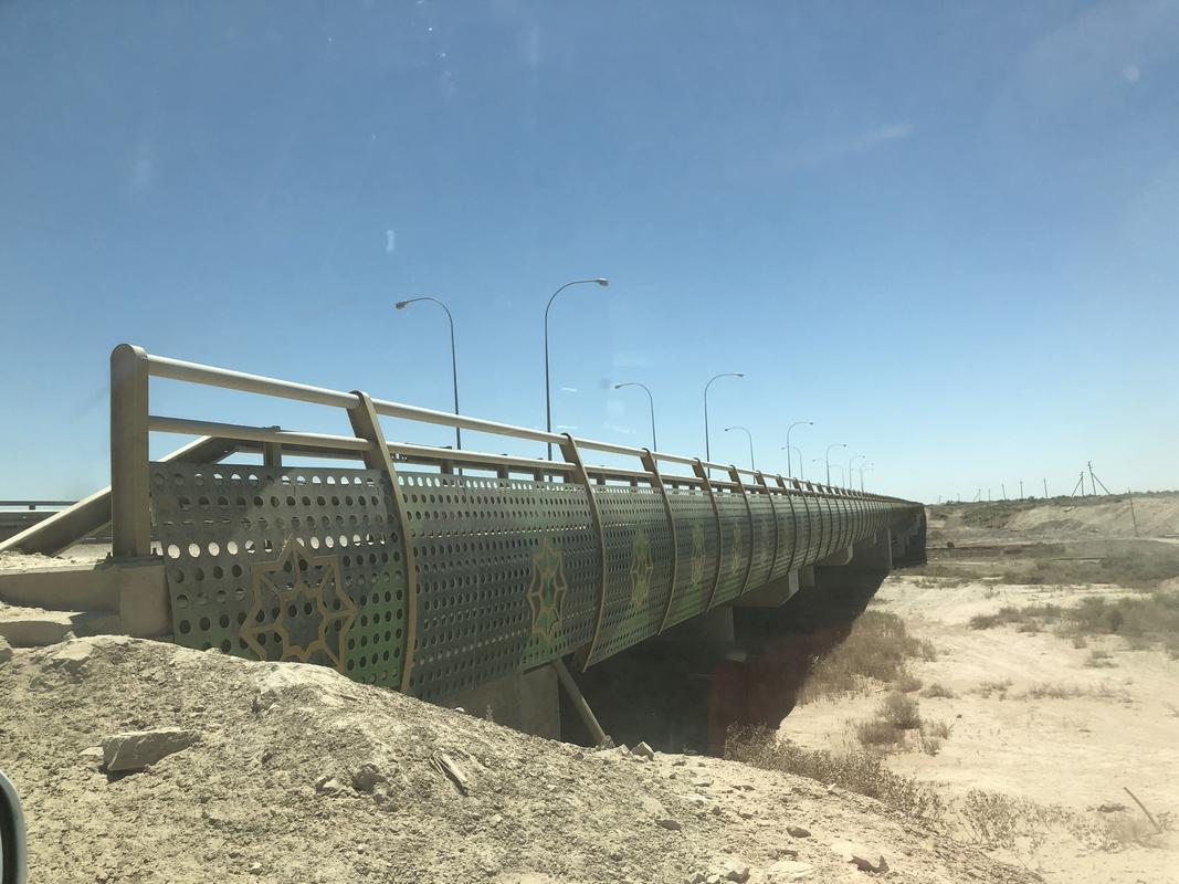 A bridge with green and gold patterned perforated railings extends over a dry, sandy landscape under a clear blue sky.