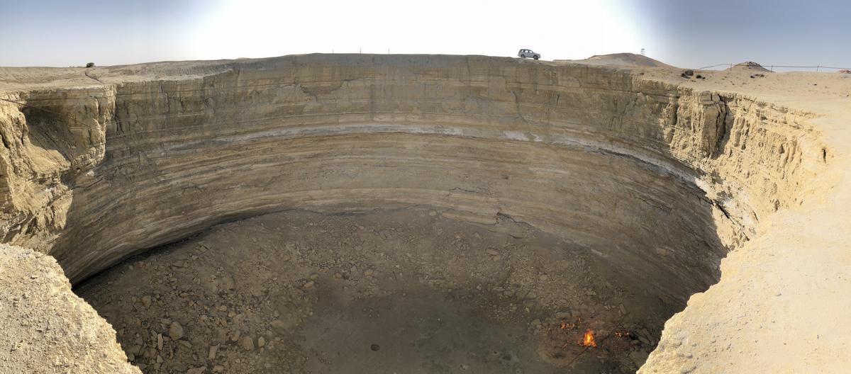 A wide panoramic view of the Darvaza Gas Crater, showing layered rock walls, a small fire at the bottom, and a vehicle on the distant rim.