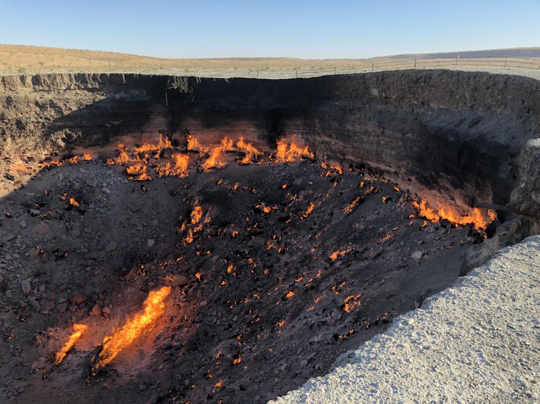 The Darvaza Gas Crater, a large pit with numerous bright orange flames burning across its dark, rocky interior in the desert.